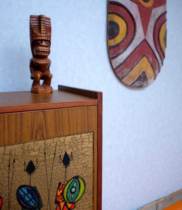 Hand Carved Wooden Tiki in the style of Lono the God of Rain on a mid centuary side bead with primitive wall art on the wall.