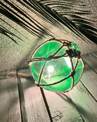 This is a photo of a Tiki Fish Float Light in green lit up and on a white washed background with tropical palm leaves