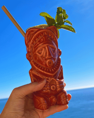 This is a photograph of a Papa New Guinean Tiki Mug, styled with a cocktail and mint garmish held against a blue sky.