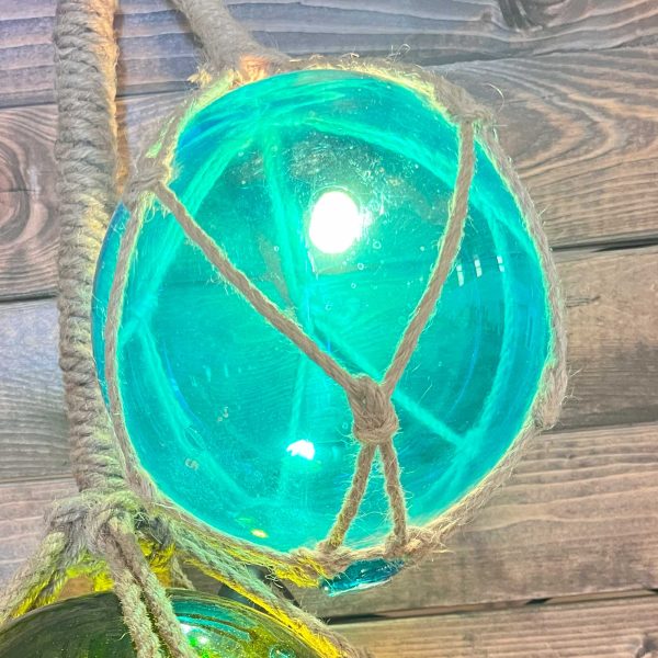 This is an image of Turquoise Glass Fish Float that has been turned into a light. It is hanging alongside another one in green and has a jute rope netting around it to hang it from.