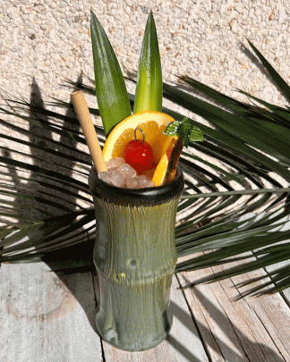 This is a picture of a ceramic Bamboo Tiki mug in green. with a black interior it is styled with a cocktail and garnish and shot on a tropical background.