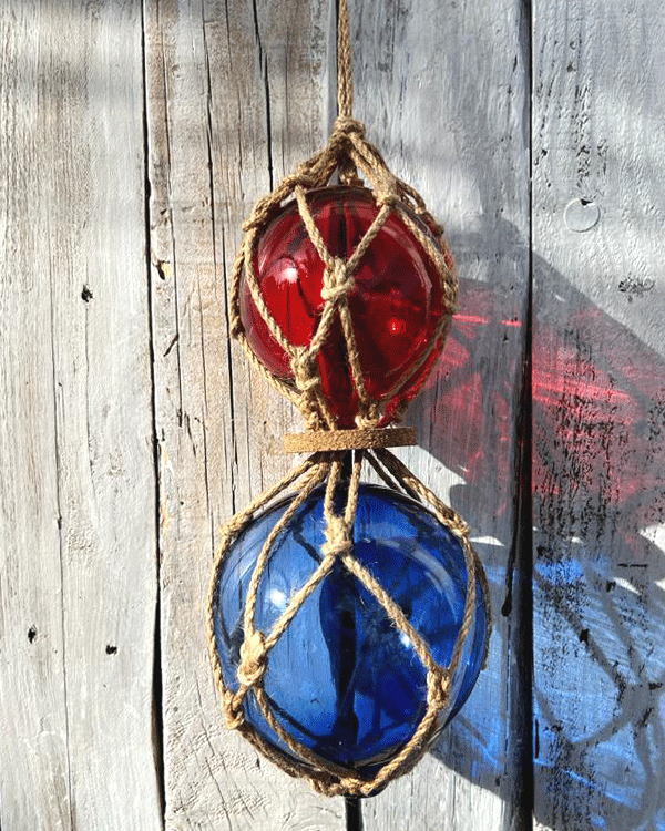 This is an image of a decorative double fish float. One red fish float with another bigger blue fish float at the bottom with rope netting on a background of white washed wood.