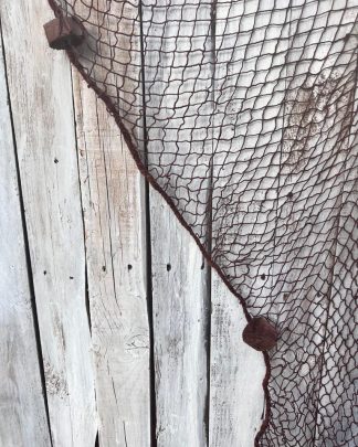 This is a photograph of a decorative cotton fish net with cork floats hanging against a distressed wooden slatted board.