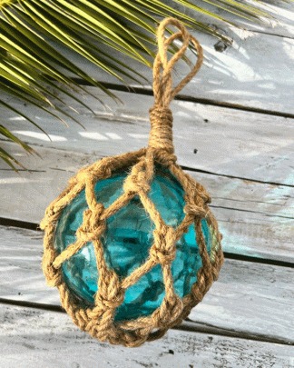 This photo is a picture of a vintage styled fish float in aqua marine with thick notted rope like a basket around it. It is shot on a whte washed wooden board with tropical foliage.