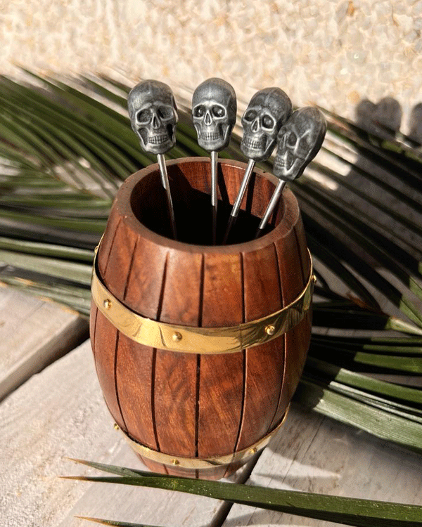 This is a picture of a Mini Tiki Barrel holding 4 Skull Cocktail picks with a tropical green palm leaf in the backgound.