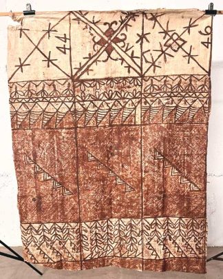 This is a photo of a piece of tapa cloth hung to show the scale of the piece. It has a traditional Tongan design painted on it in brown and creams