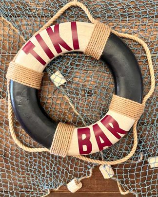 This is an image of a replica life ring in blue and cream with the words Tiki Bar written on it. The life ring is decorated with tradition jute rope which create the hanging loops. It is shot against some fishing nets.